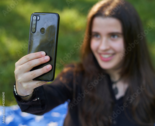 young woman with long hair in a black shirt and white pants sitting on a blanket of stars on the grass in a park with her bag using her phone to take pictures and video conferencing. influencer nature