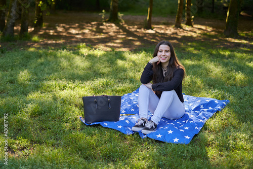 Pretty young woman with long hair in a black shirt and white pants sitting on a blanket of stars on the grass in a park with her bag looking to the sides. teenager resting in nature with blue blanket