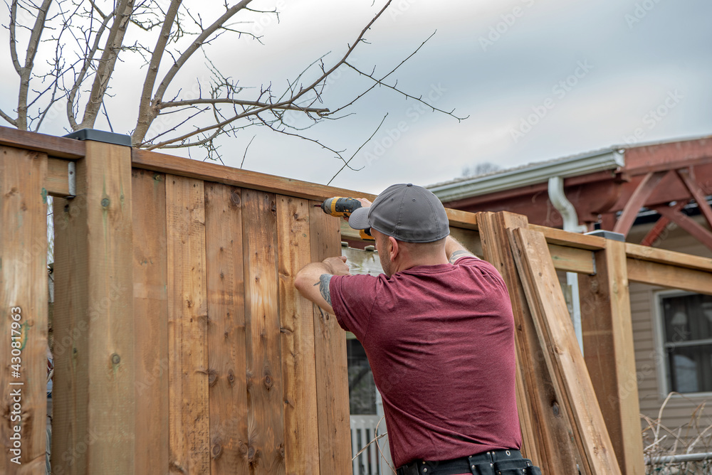 A white, middle-aged gay man builds a wooden fence in his back yard.