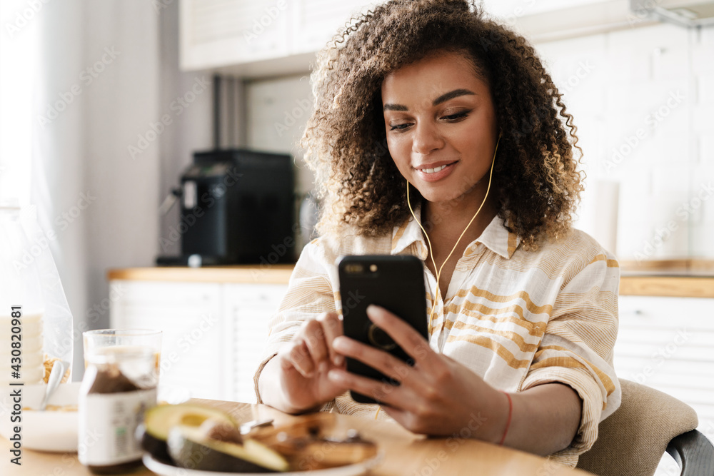 The smiling woman in the wired headphones looking at the phone while sitting at the table in the kitchen The smiling woman in the wired headphones looking at the phone while sitting at the table in the kitchen