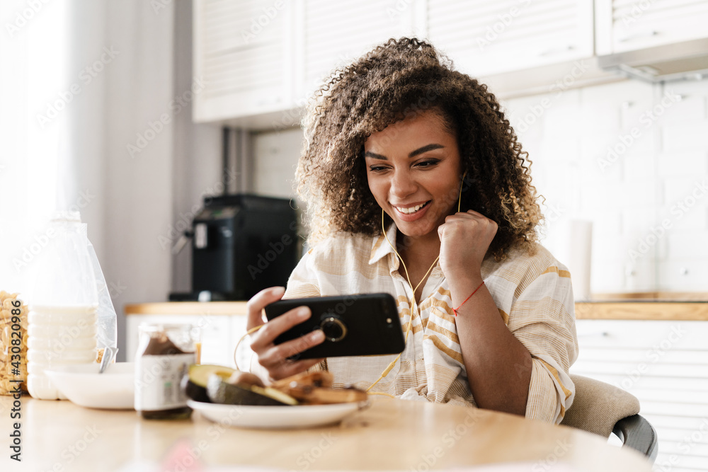The laughing woman in the wired headphones looking at the phone while sitting at the table in the kitchen The laughing woman in the wired headphones looking at the phone while sitting at the table in the kitchen