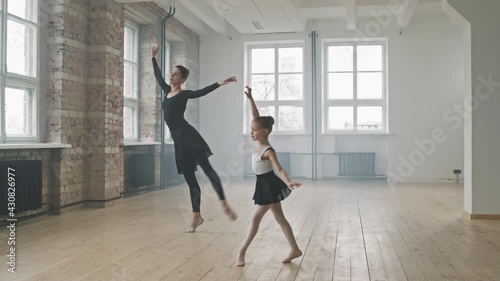 Full shot of young professional ballerina in black ballet dress dancing together with pretty little girl, moving synchronically at dance class photo