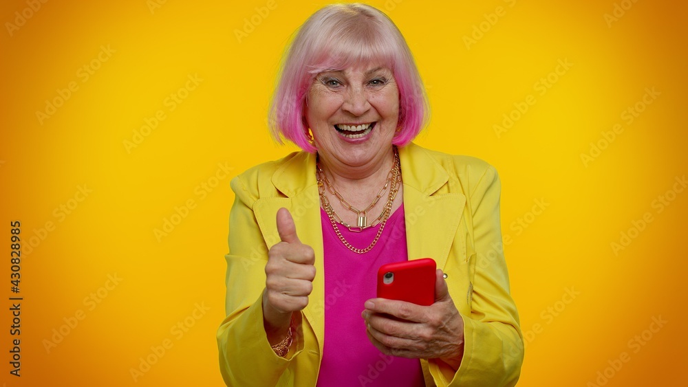 Happy excited joyful elderly granny gray-haired woman use mobile cell phone typing browsing say wow yes, found out great big win news doing winner gesture. Senior old grandmother on orange background Happy excited joyful elderly granny gray-haired woman use mobile cell phone typing browsing say wow yes, found out great big win news doing winner gesture. Senior old grandmother on orange background