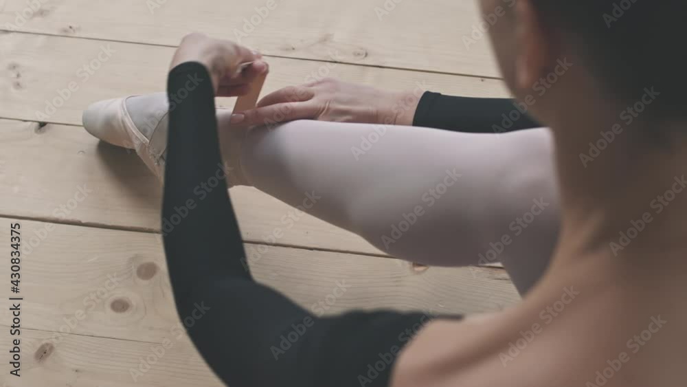 Over-the-shoulder shot of gracious young ballerina tying her pointe shoes before ballet dancing in choreography studio Over-the-shoulder shot of gracious young ballerina tying her pointe shoes before ballet dancing in choreography studio