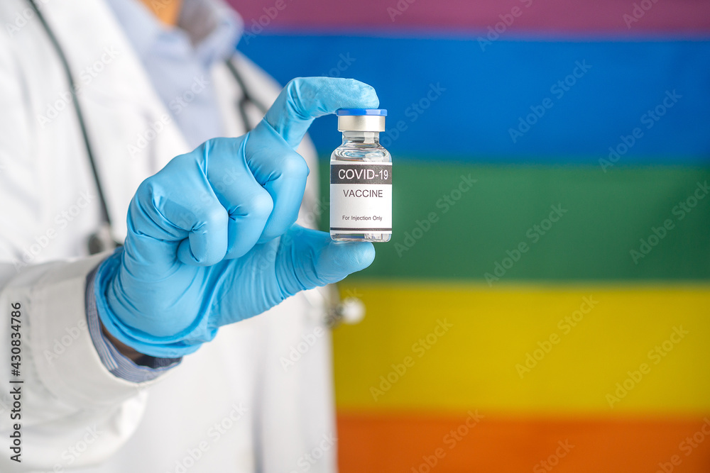 A doctor holding a COVID-19 vaccine bottle prepares vaccination against the background of the rainbow flag (LGBT). Vaccine for immunization and treatment from virus infection. Medical concept A doctor holding a COVID-19 vaccine bottle prepares vaccination against the background of the rainbow flag (LGBT). Vaccine for immunization and treatment from virus infection. Medical concept