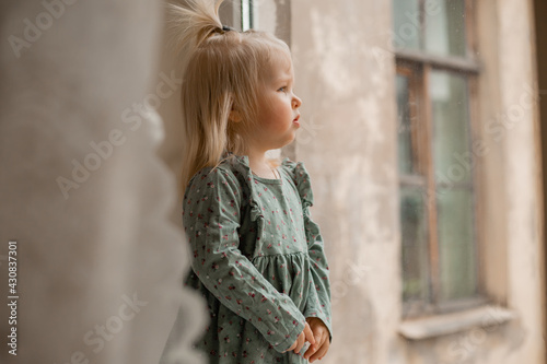 Cute baby blonde girl in vintage dress stands by the window of the house