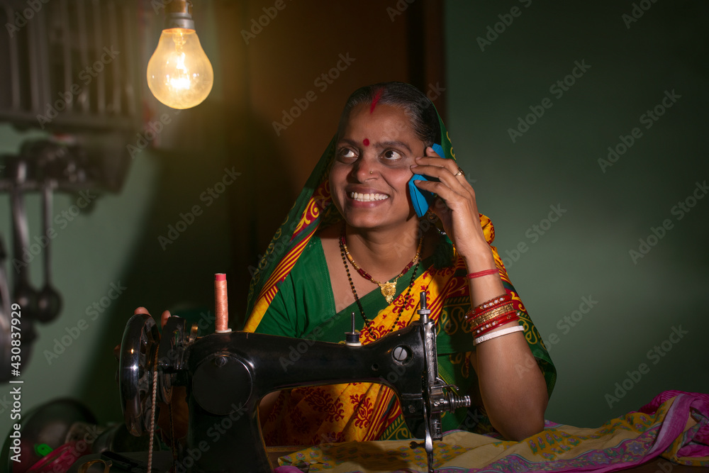 Indian rural woman using sewing machine and talking on a mobile phone Indian rural woman using sewing machine and talking on a mobile phone