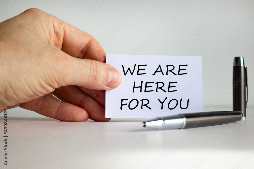 Help and support symbol. White paper with words 'we are here for you' in businessman hand, metalic pen. Beautiful white background. Business, help and support concept. Copy space. Help and support symbol. White paper with words 'we are here for you' in businessman hand, metalic pen. Beautiful white background. Business, help and support concept. Copy space.