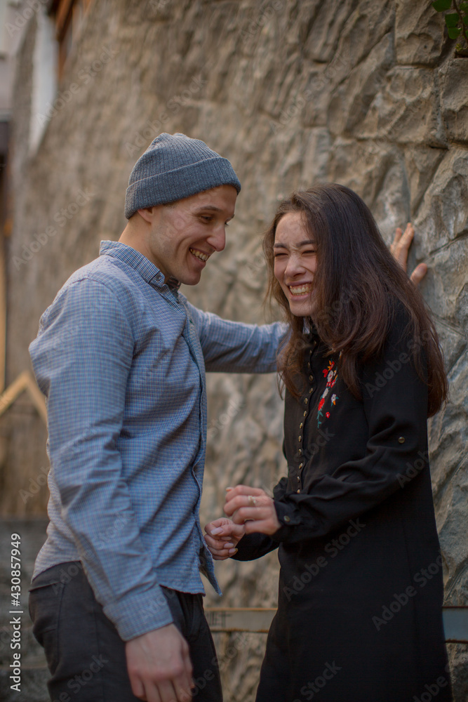 A young couple in love are smiling at each other while standing at a stone wall in the old city in clear weather. Love story. Young happy couple. High quality photo