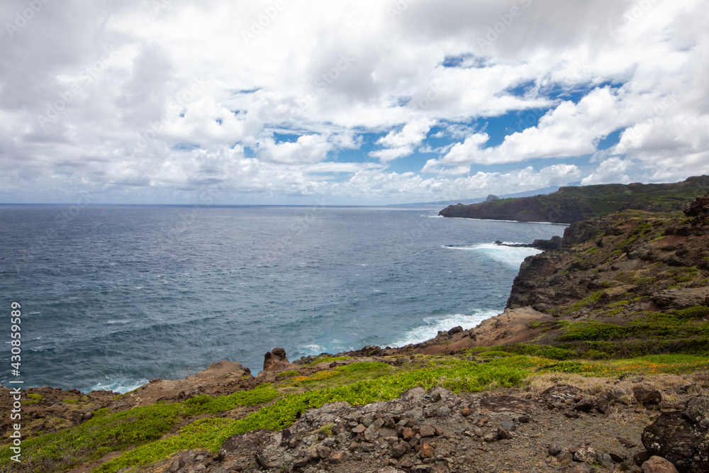 Wide landscape view of a rocky and grassy coastline with cloudy blue sky and deep blue ocean in Maui, Hawaii.