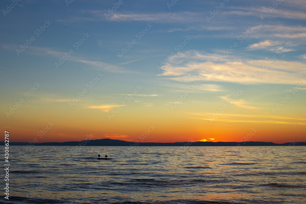 Scenic sunset on lake in dusk with blue clear sky and cirrus clouds, gulls on stone in calm waves under sundown blazing