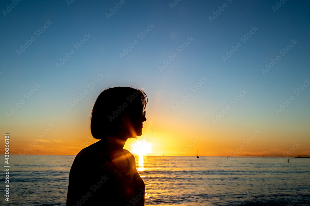 The silhouette of a young woman wearing sunglasses is seen against a sunset sky with the Pacific Ocean in the background. 