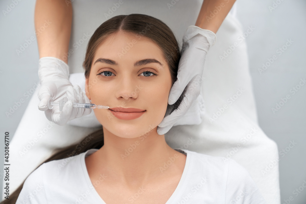 Top view portrait of smiling attractive woman lying in beautician and doing injection in lip with special preparation. Concept of process procedure lip augmentation in professional salon. Top view portrait of smiling attractive woman lying in beautician and doing injection in lip with special preparation. Concept of process procedure lip augmentation in professional salon.