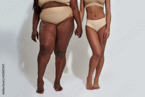 Legs of two african american women in underwear with different body size holding each other hands, standing together isolated over gray background