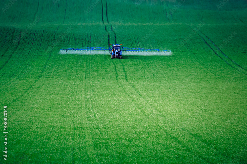 Obraz premium A tractor with attachments performs agricultural work in a green field. Business in industrial agriculture, mechanized processing on the farm. Selective focus, natural background.
