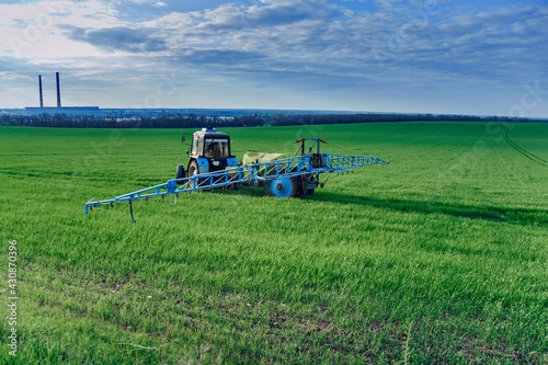 A tractor with attachments performs agricultural work in a green field. Business in industrial agriculture, mechanized processing on the farm. Selective focus, natural background.