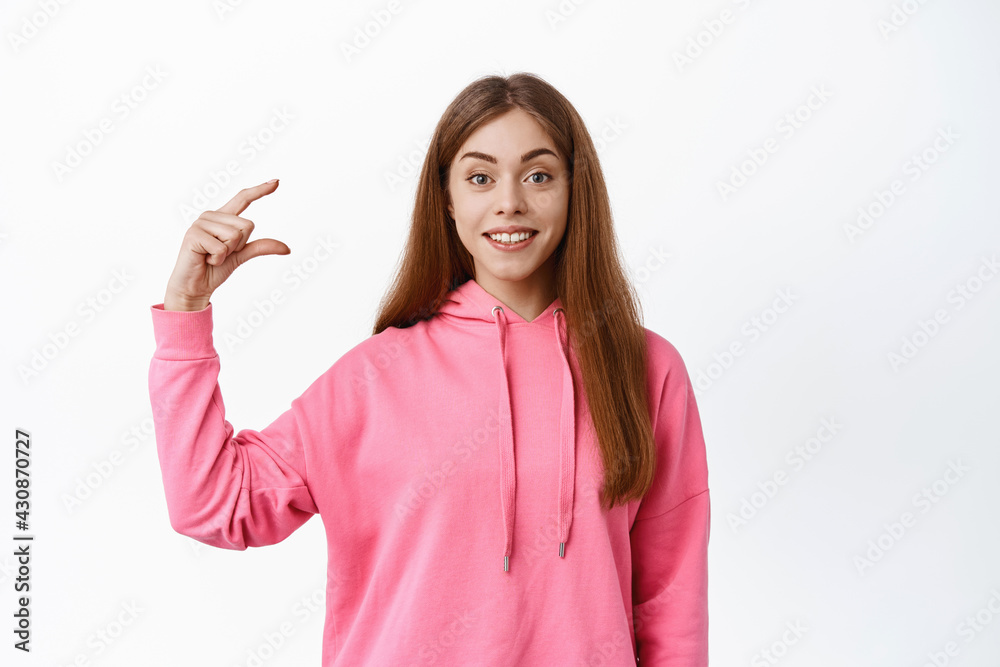 Young woman showing little size, small thing gesture and smiling, display tiny item, standing over white background Young woman showing little size, small thing gesture and smiling, display tiny item, standing over white background