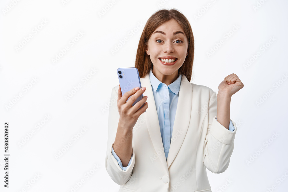 Portrait of lucky office woman in business suit, jumping from triumph and excitement, holding smartphone, rejoicing and saying yes, winning online, white background Portrait of lucky office woman in business suit, jumping from triumph and excitement, holding smartphone, rejoicing and saying yes, winning online, white background