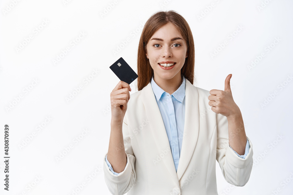 Confident, successful business woman shows plastic credit card and thumbs up, smiling pleased, recommend bank, standing over white background Confident, successful business woman shows plastic credit card and thumbs up, smiling pleased, recommend bank, standing over white background