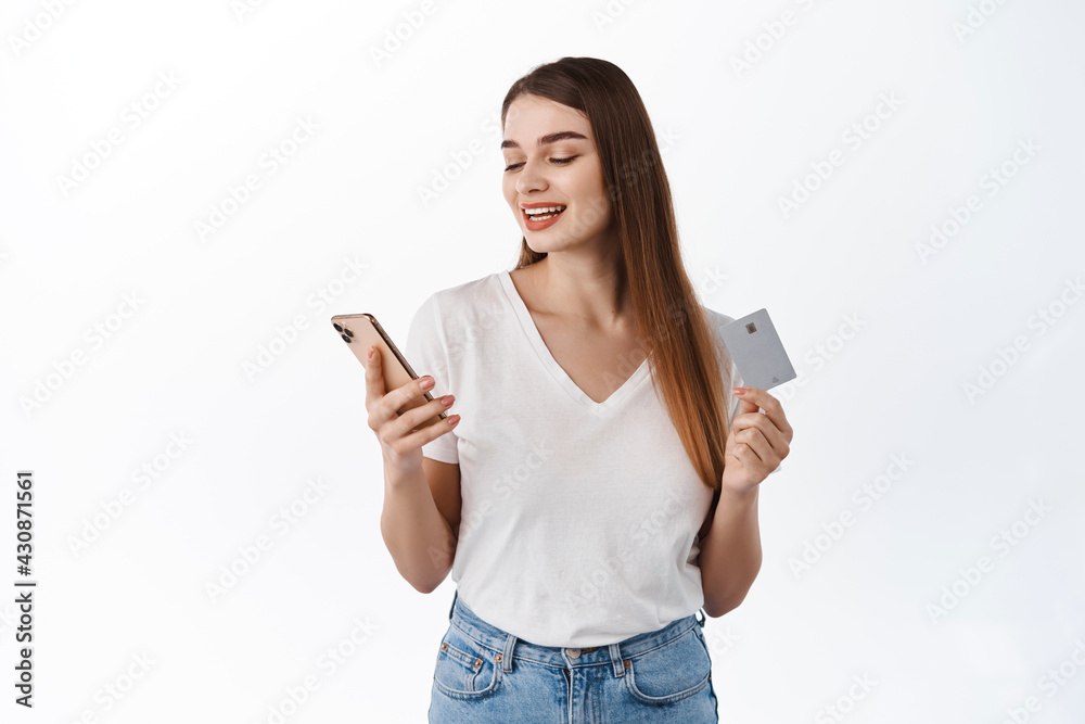 Contactless payments and online delivery concept. Young woman looks at her phone while paying with credit card, using shopping app or making transaction, standing over white background Contactless payments and online delivery concept. Young woman looks at her phone while paying with credit card, using shopping app or making transaction, standing over white background