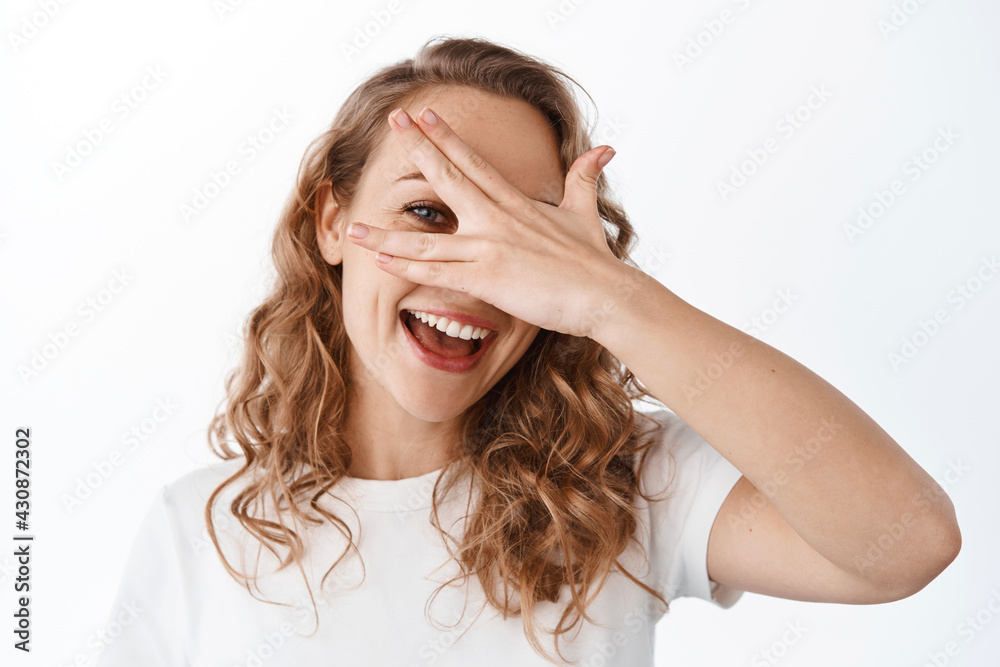 Carefree beautiful woman look through fingers, covering eyes with hand and smiling happy, standing in t-shirt against white background Carefree beautiful woman look through fingers, covering eyes with hand and smiling happy, standing in t-shirt against white background