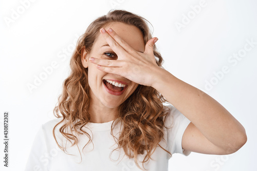 Carefree beautiful woman look through fingers, covering eyes with hand and smiling happy, standing in t-shirt against white background