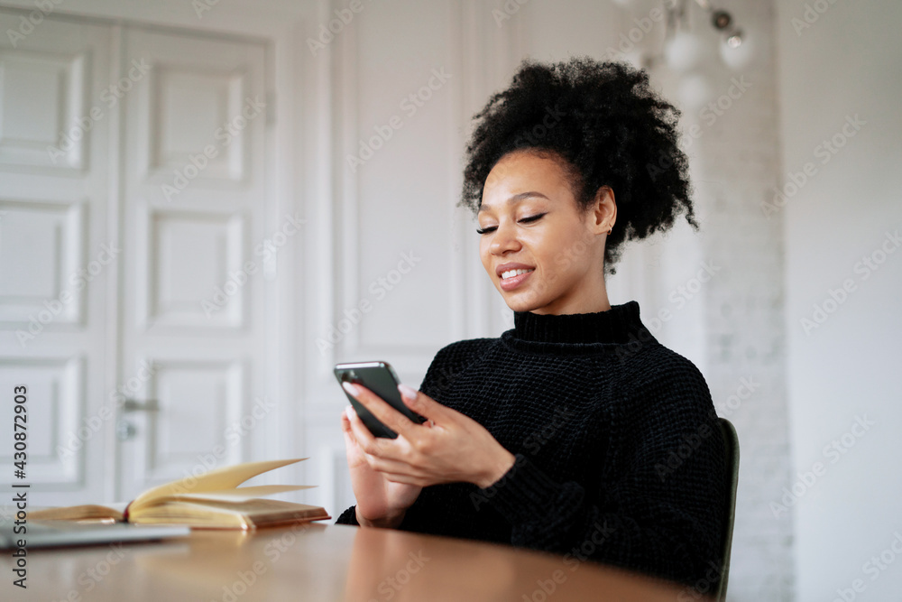 Prints a message on the phone in the messenger. A satisfied female student designer smiles at an Afro-ethnic appearance. In the browser, working on a new work project. Curly black hair. Prints a message on the phone in the messenger. A satisfied female student designer smiles at an Afro-ethnic appearance. In the browser, working on a new work project. Curly black hair.