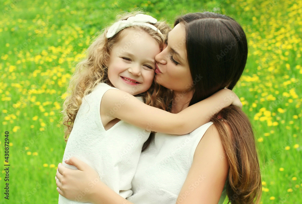 Portrait of little child daughter kissing her happy smiling mother on the grass in a summer day Portrait of little child daughter kissing her happy smiling mother on the grass in a summer day