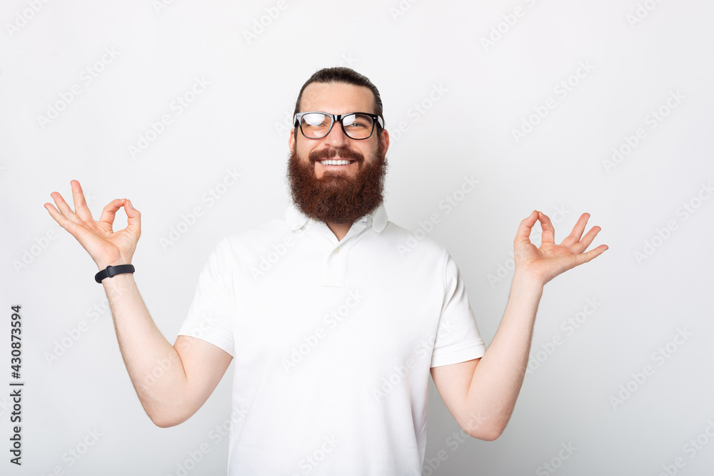 A calm smiling man is looking at the camera happy in a zen pose near a white wall A calm smiling man is looking at the camera happy in a zen pose near a white wall
