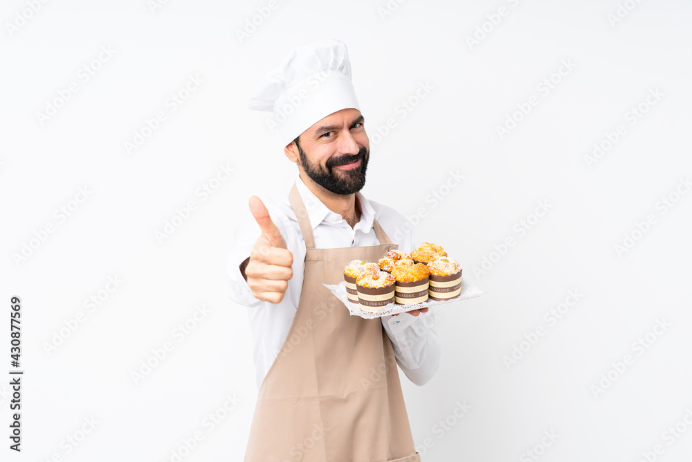 Young man holding muffin cake over isolated white background with thumbs up because something good has happened Young man holding muffin cake over isolated white background with thumbs up because something good has happened