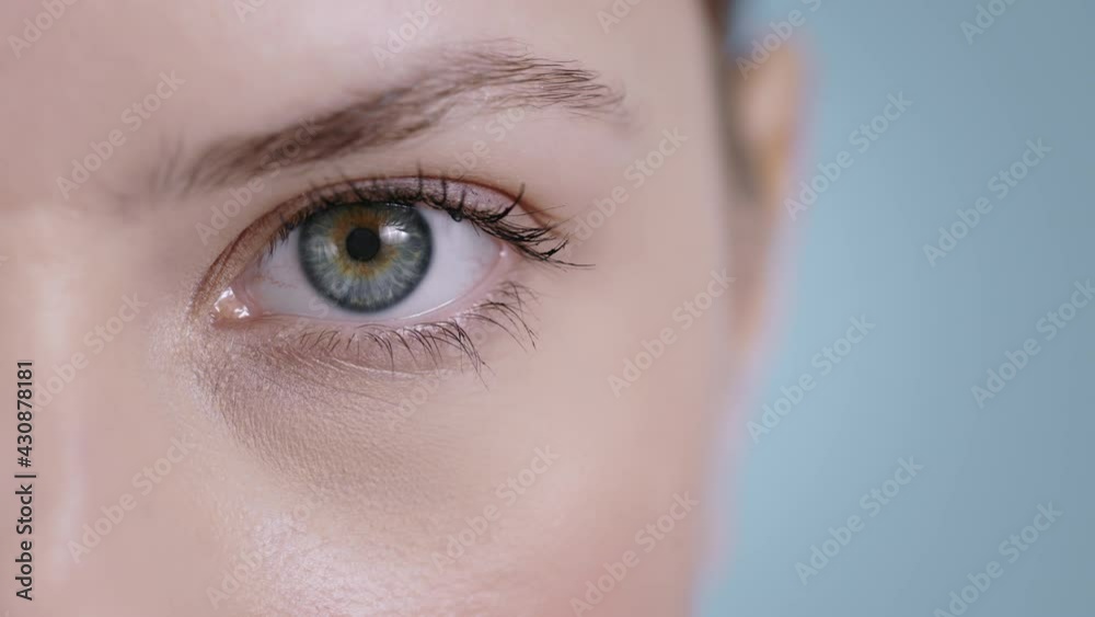 Close up view of attractive young woman face half opening eye and looking to camera . Portrait of pretty female person with light nude make up posing at studio background.