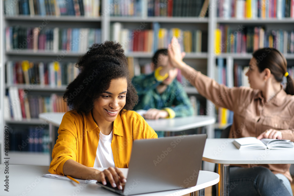 Happy confident smart african american teenage girl sitting at table in library, using laptop while preparing for lessons or exam, two blurry students give each other five in the background Happy confident smart african american teenage girl sitting at table in library, using laptop while preparing for lessons or exam, two blurry students give each other five in the background