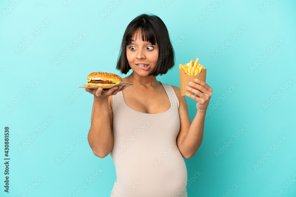 Young pregnant woman holding burger and fried chips over isolated blue background Young pregnant woman holding burger and fried chips over isolated blue background