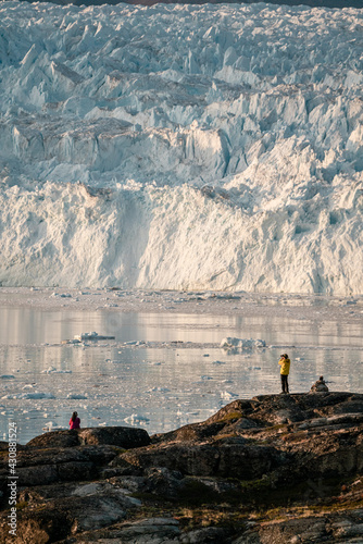 Unidentified People sitting standing in front of huge glacier wall of ice. Eqip Sermia Glacier Eqi glacier in Greenland called the calving glacier during midnight sun. Hikers during travel and photo