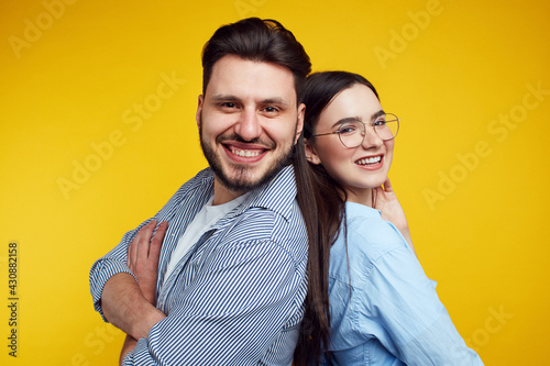 Couple stands backs and smiling against yellow background