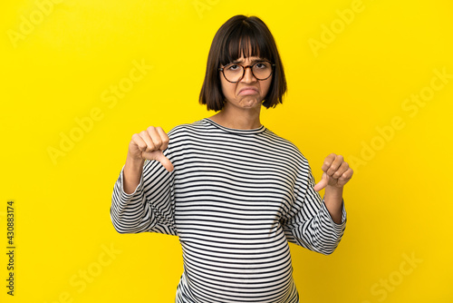 Young pregnant woman over isolated yellow background showing thumb down with two hands