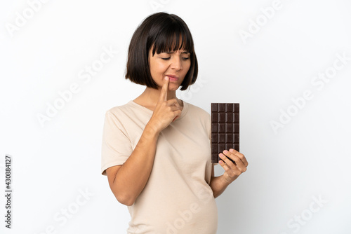 Young mixed race pregnant woman isolated on white background taking a chocolate tablet and having doubts