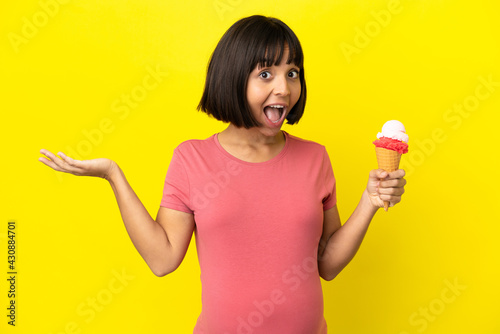 Pregnant woman holding a cornet ice cream isolated on yellow background with shocked facial expression © luismolinero