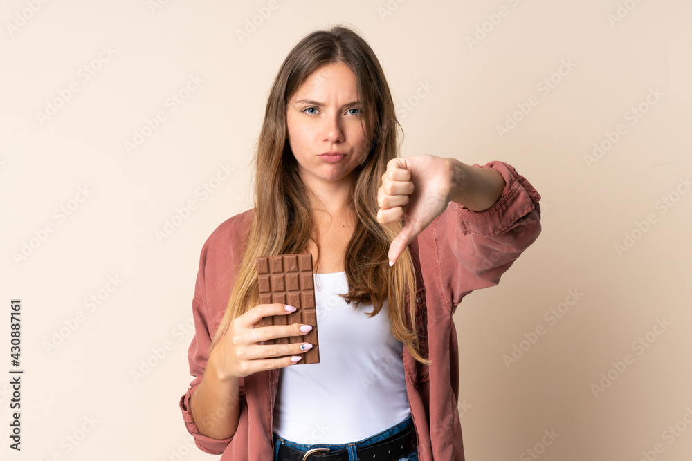 Young Lithuanian woman isolated on beige background taking a chocolate tablet making bad signal Young Lithuanian woman isolated on beige background taking a chocolate tablet making bad signal
