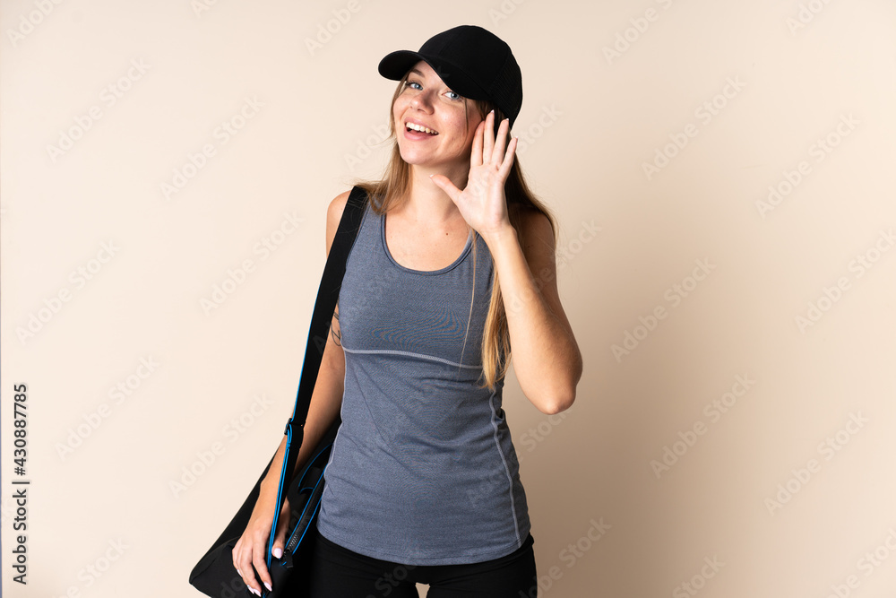 Young sport Lithuanian woman holding a sport bag isolated on beige background listening to something by putting hand on the ear Young sport Lithuanian woman holding a sport bag isolated on beige background listening to something by putting hand on the ear