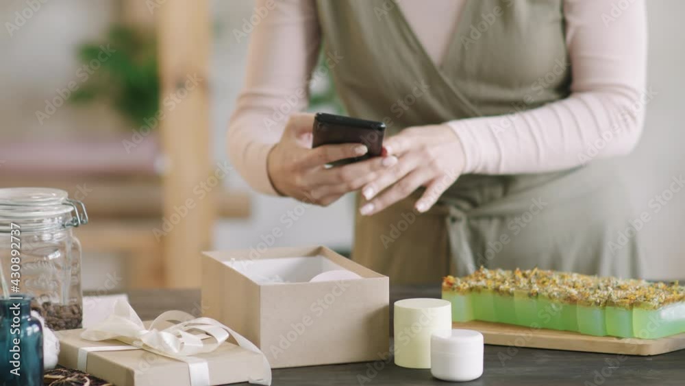 Midsection PAN shot of unrecognizable woman taking photos of handmade natural cosmetics – creams and soap – on her smartphone before posting