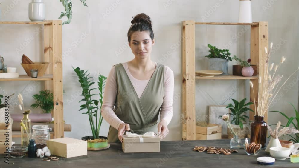 Medium PAN shot of young attractive woman packing handmade cosmetic products – face cream and soap – into beautiful present box then looking at camera smiling