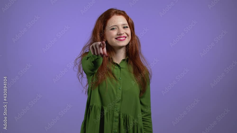 Charming woman points index finger into camera lens and winks, flirts. Hey, you. Pretty Girl on violet background. Charming woman points index finger into camera lens and winks, flirts. Hey, you. Pretty Girl on violet background.