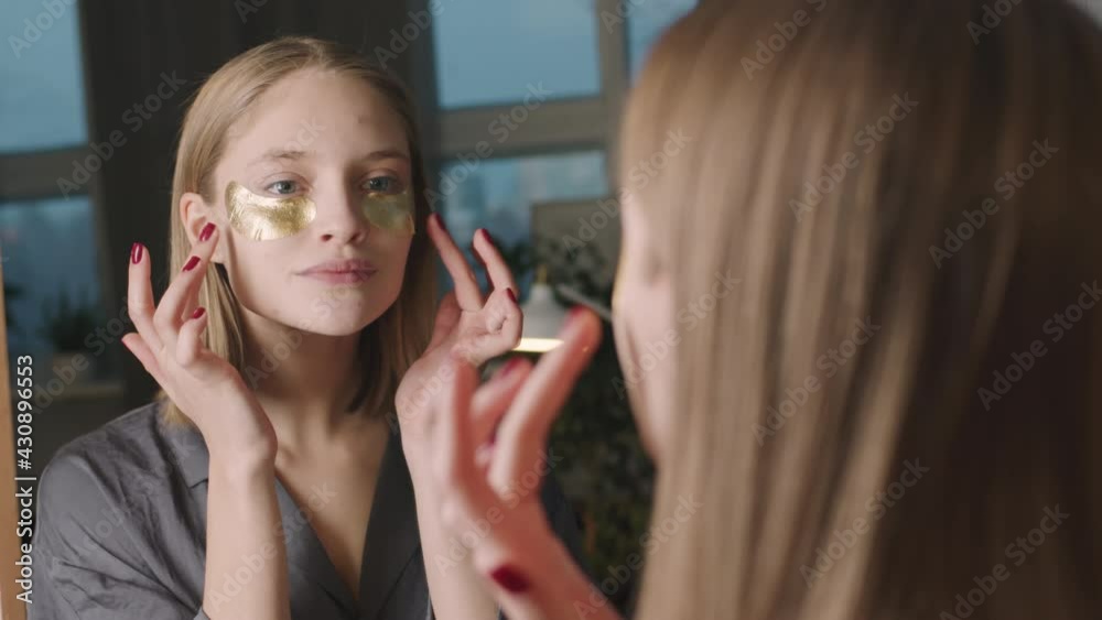 Medium shot of young natural woman applying golden under-eye patches on face looking in mirror doing evening beauty routine Medium shot of young natural woman applying golden under-eye patches on face looking in mirror doing evening beauty routine