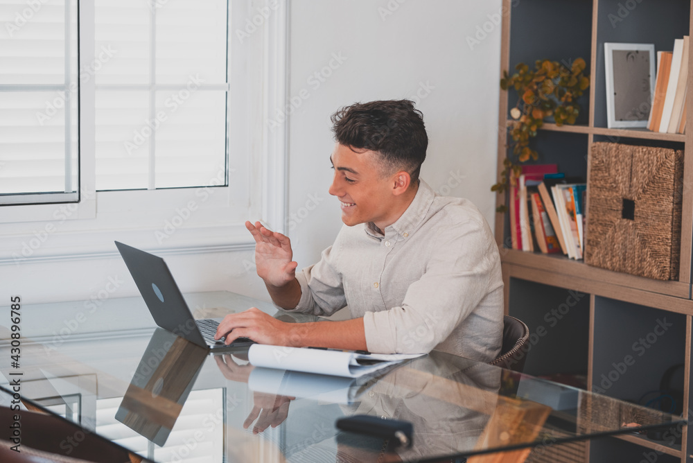 Happy young man teenager smiling and talking in video conference studying and learning online with school. Millennial doing homework at home calling . Happy young man teenager smiling and talking in video conference studying and learning online with school. Millennial doing homework at home calling .