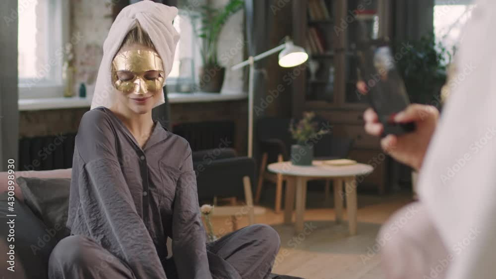 Medium shot of young smiling woman in facial golden sheet mask posing to smartphone camera having fun with her female friend spending weekend at home doing beauty skincare procedures Medium shot of young smiling woman in facial golden sheet mask posing to smartphone camera having fun with her female friend spending weekend at home doing beauty skincare procedures