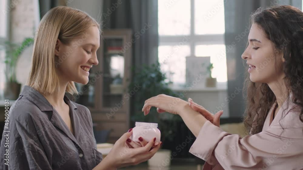 PAN waist-up shot with slowmo of young beautiful brunette woman applying some strawberry body butter on her hands sitting in front of female friend doing beauty routine together PAN waist-up shot with slowmo of young beautiful brunette woman applying some strawberry body butter on her hands sitting in front of female friend doing beauty routine together