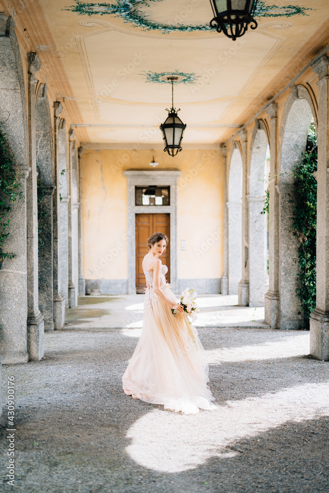 Bride in a beautiful dress with a bouquet of pink flowers walks through the vaulted hall. Lake Como, Italy. Back view