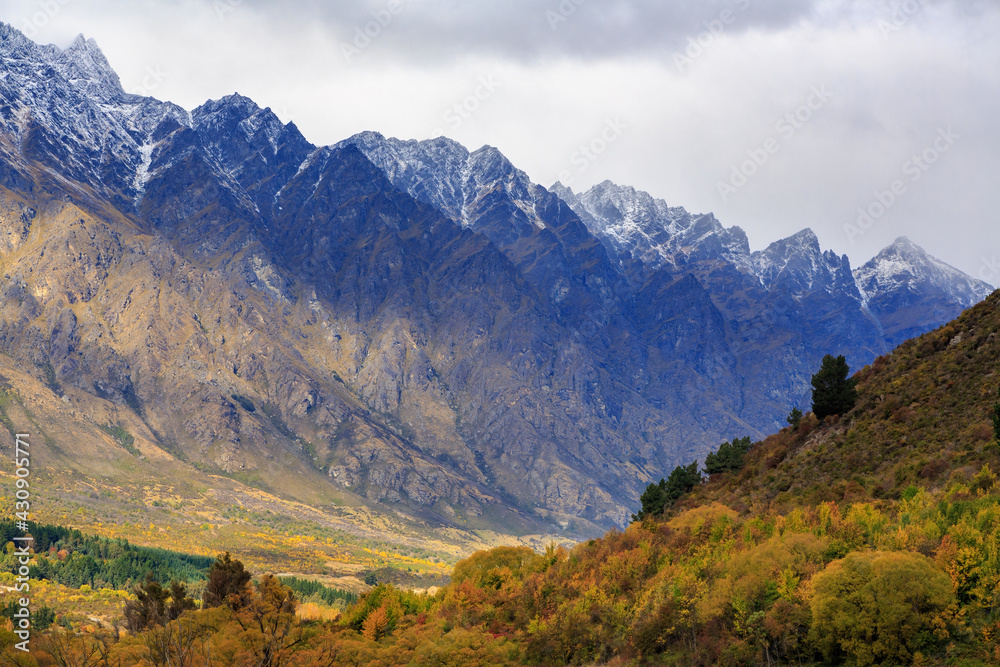 The Remarkables mountain range near Queenstown, New Zealand, with a dusting of snow on the highest slopes. In the foreground are trees with autumn foliage