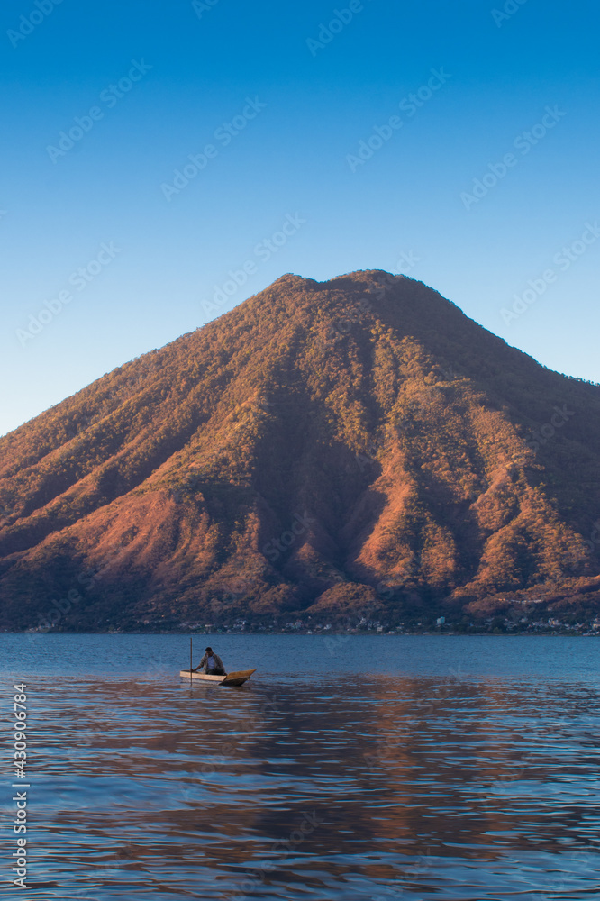 Obraz premium man sailing in a wooden boat on a lake in the middle of a beautiful sunrise with a dormant volcano in the background